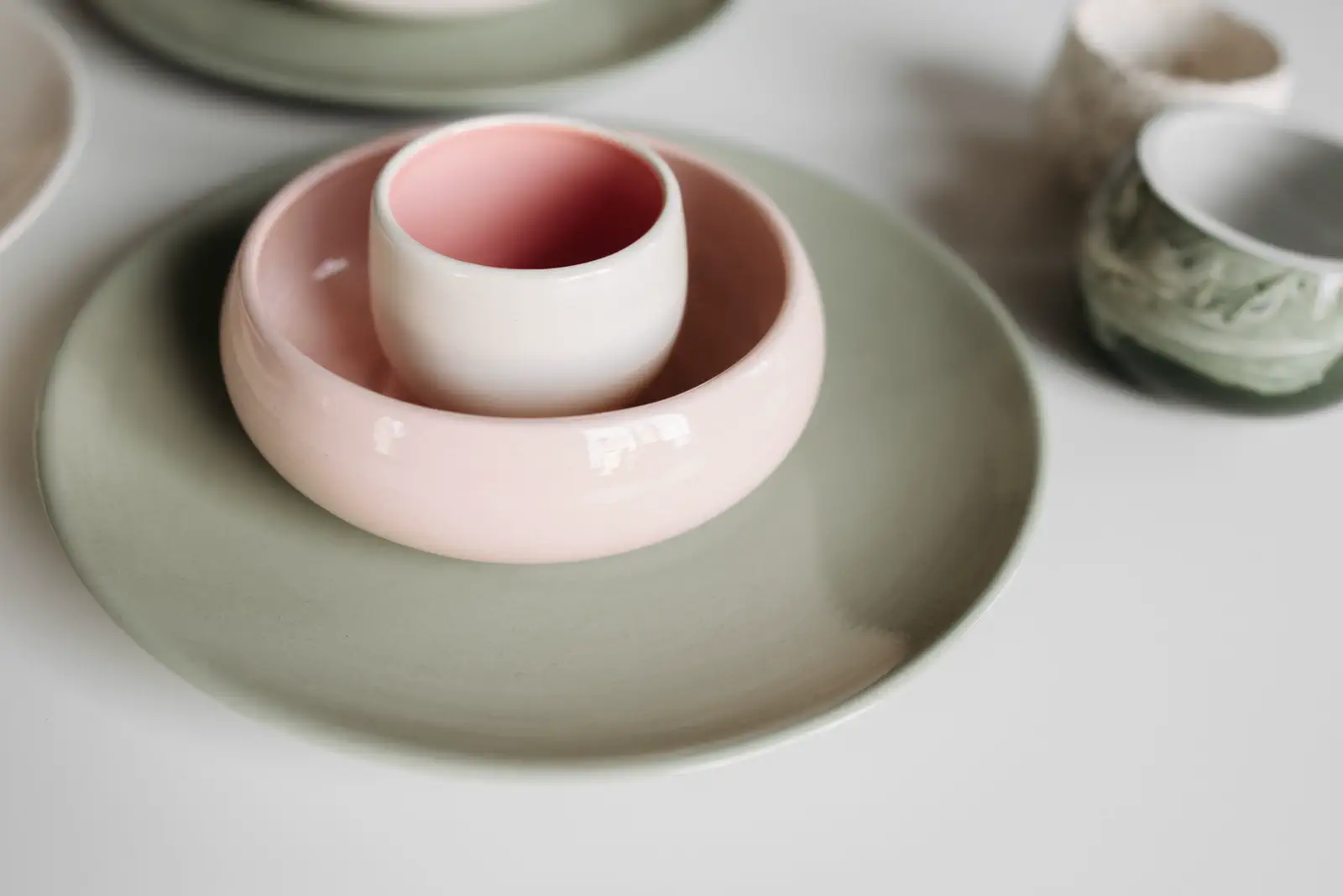 A pink bowl and cup on a sage-green plate on a white table, more cups behind.