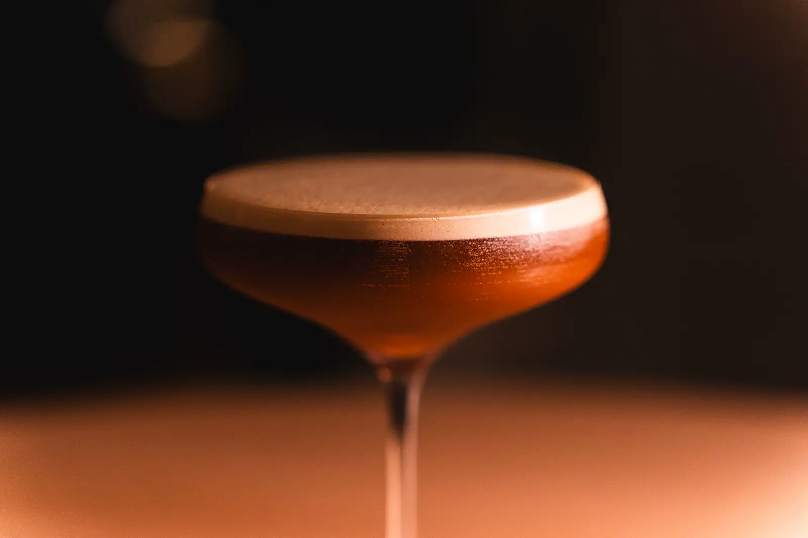 A frothed coupe cocktail in a narrow pool of warm light.