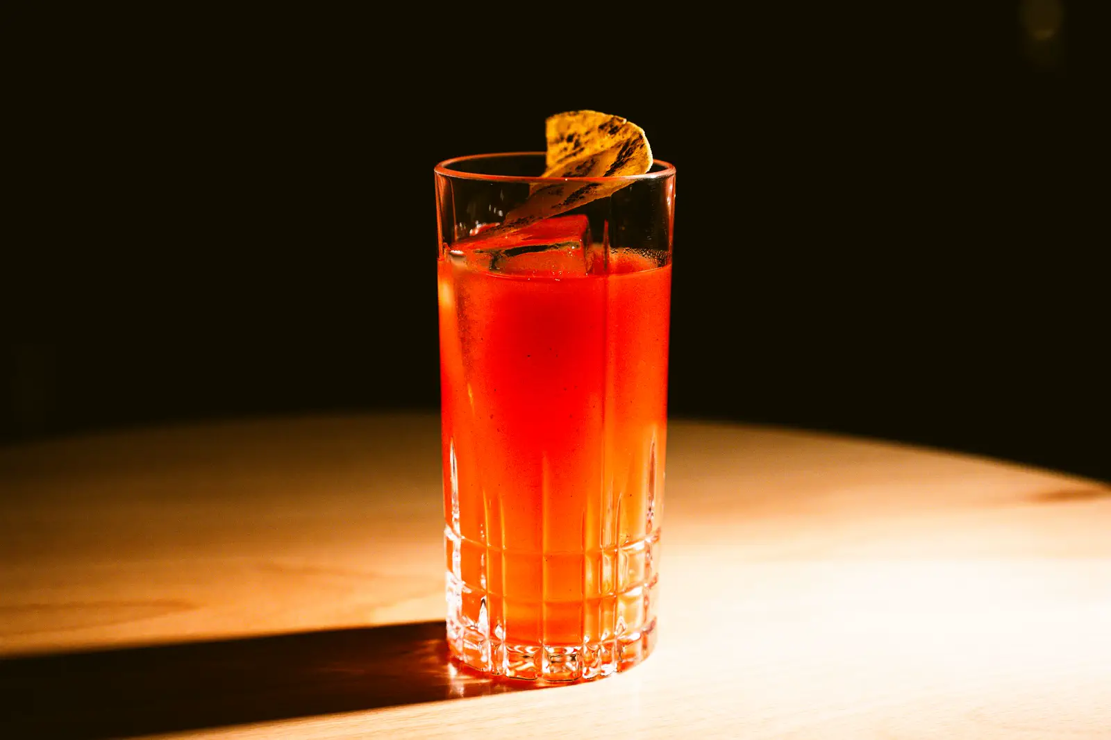 A tall red cocktail with a dried citrus wheel in a cut-glass highball on a pale wooden table.
