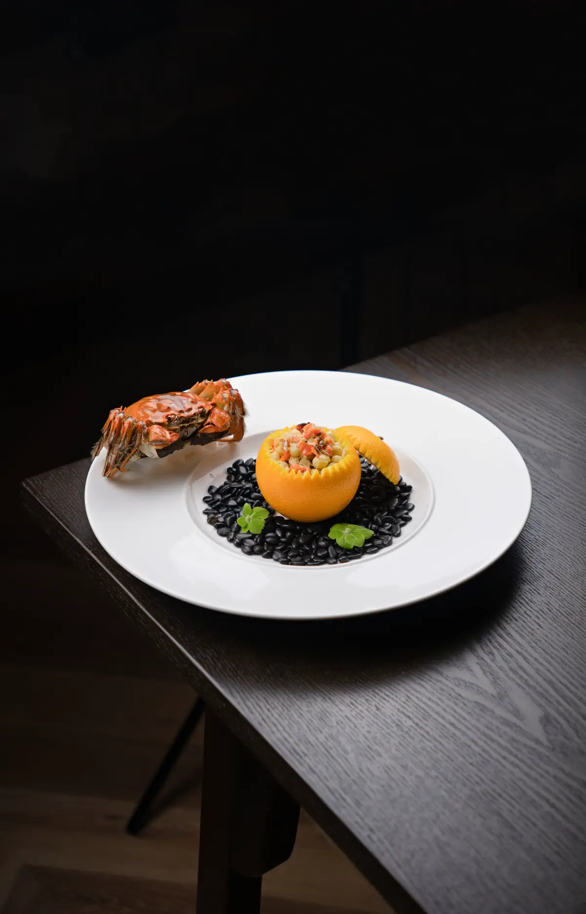 A whole crab beside a hollowed orange filled with diced vegetables on black rice, on a white plate on a dark wood table.
