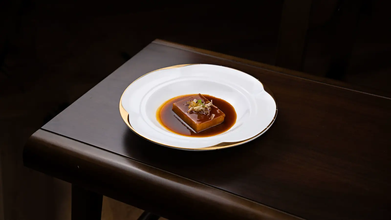 A braised cube in dark glossy sauce on a scalloped white dish on the corner of a dark wood table.