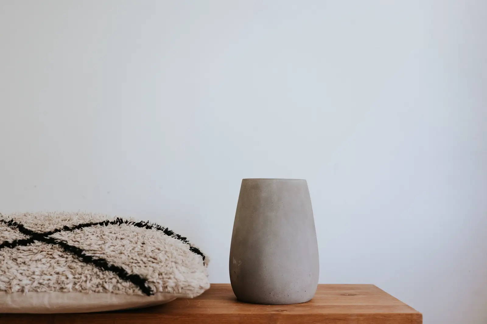 A grey ceramic vase and a shaggy wool cushion on a wooden bench against a white wall.