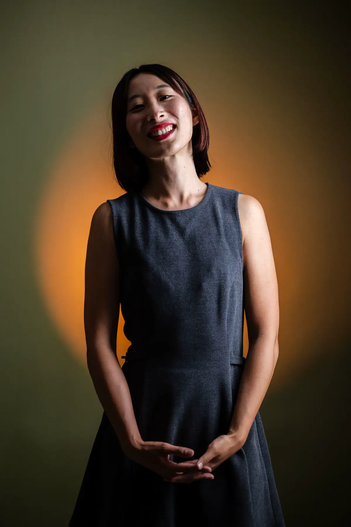 The woman smiling with her hands clasped, lit from the front against the warm glow.