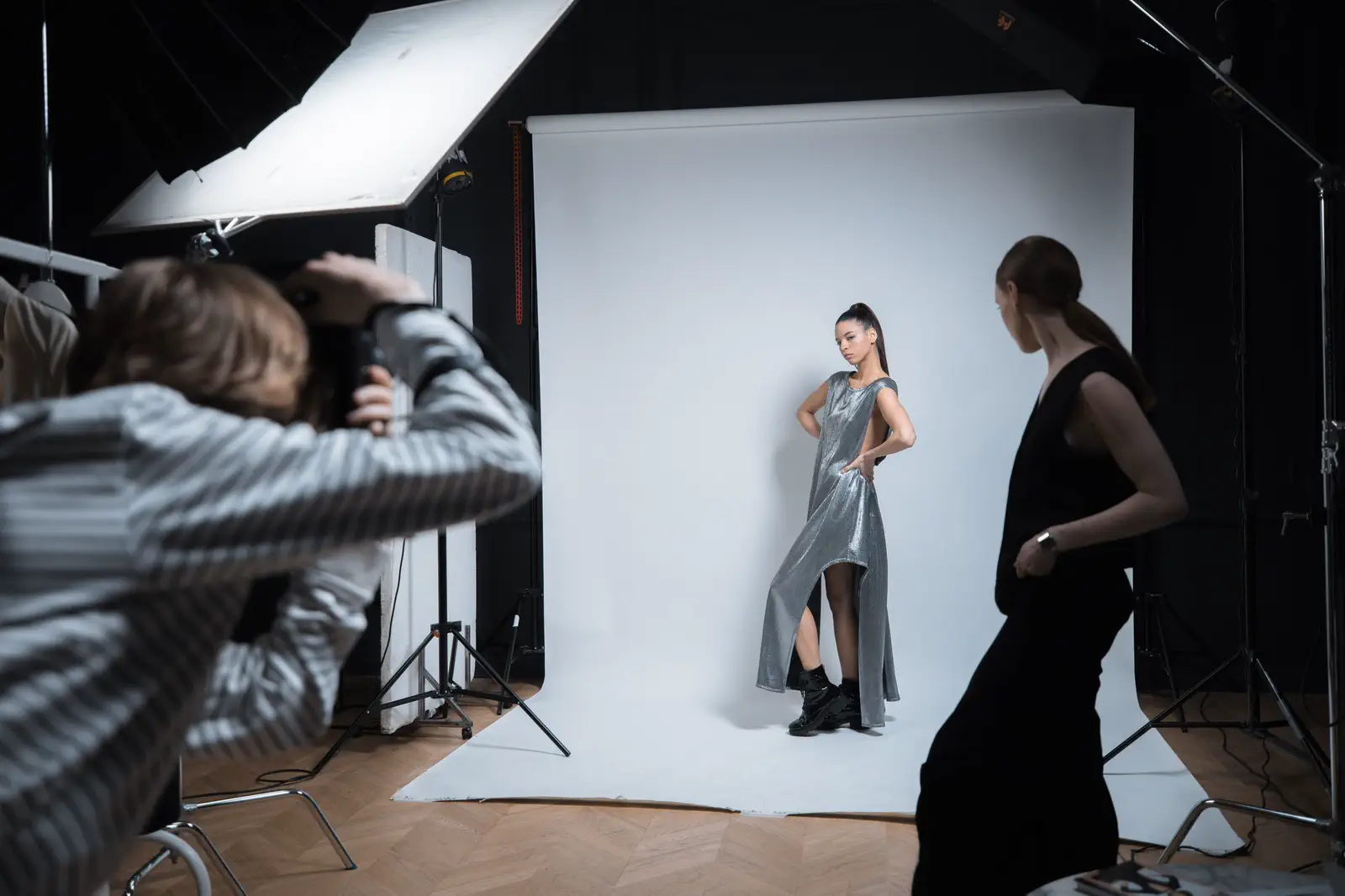 A photographer shooting a model in a silver dress on white paper while a stylist waits.