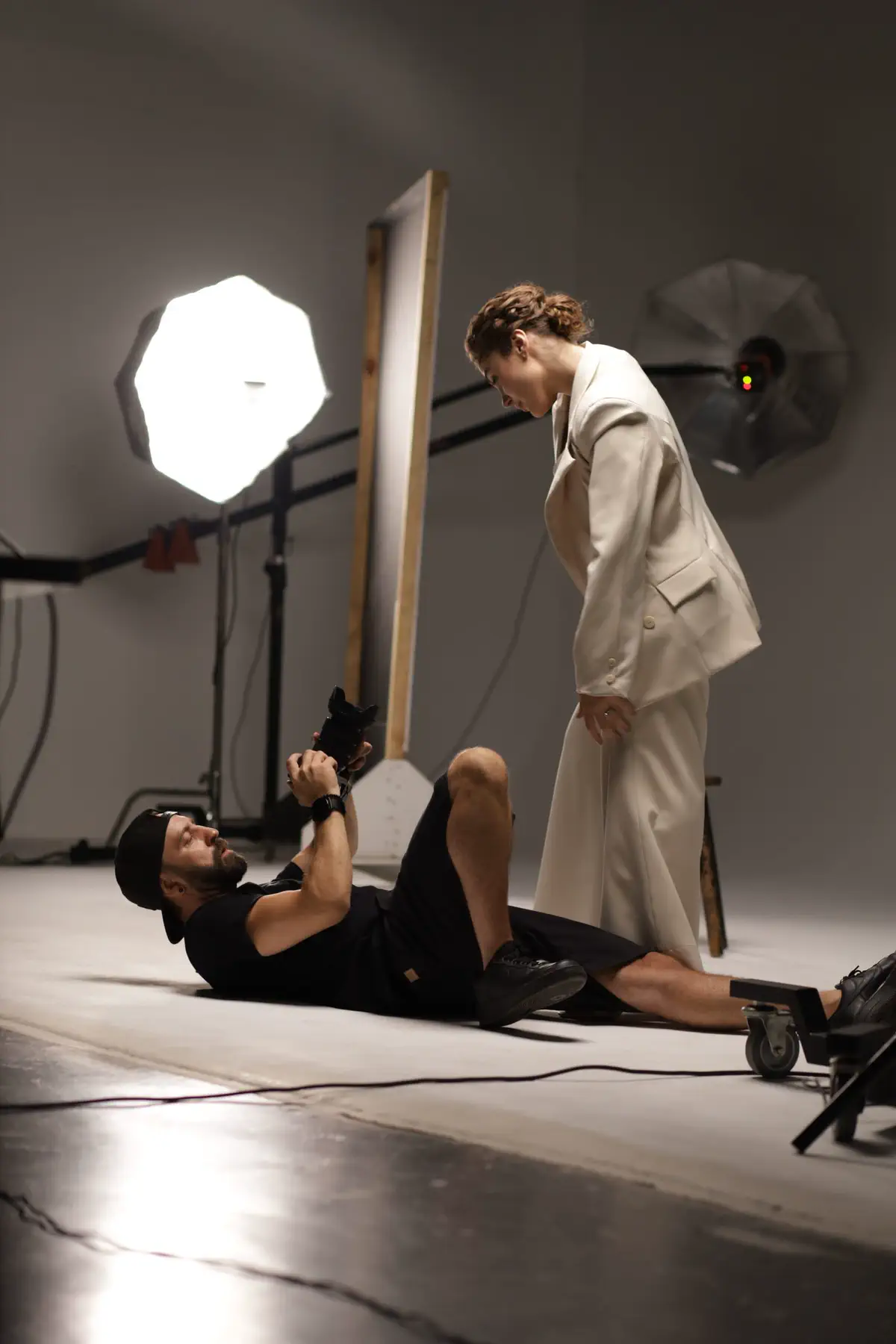 A photographer lying on the studio floor shooting up at a model in a cream suit.