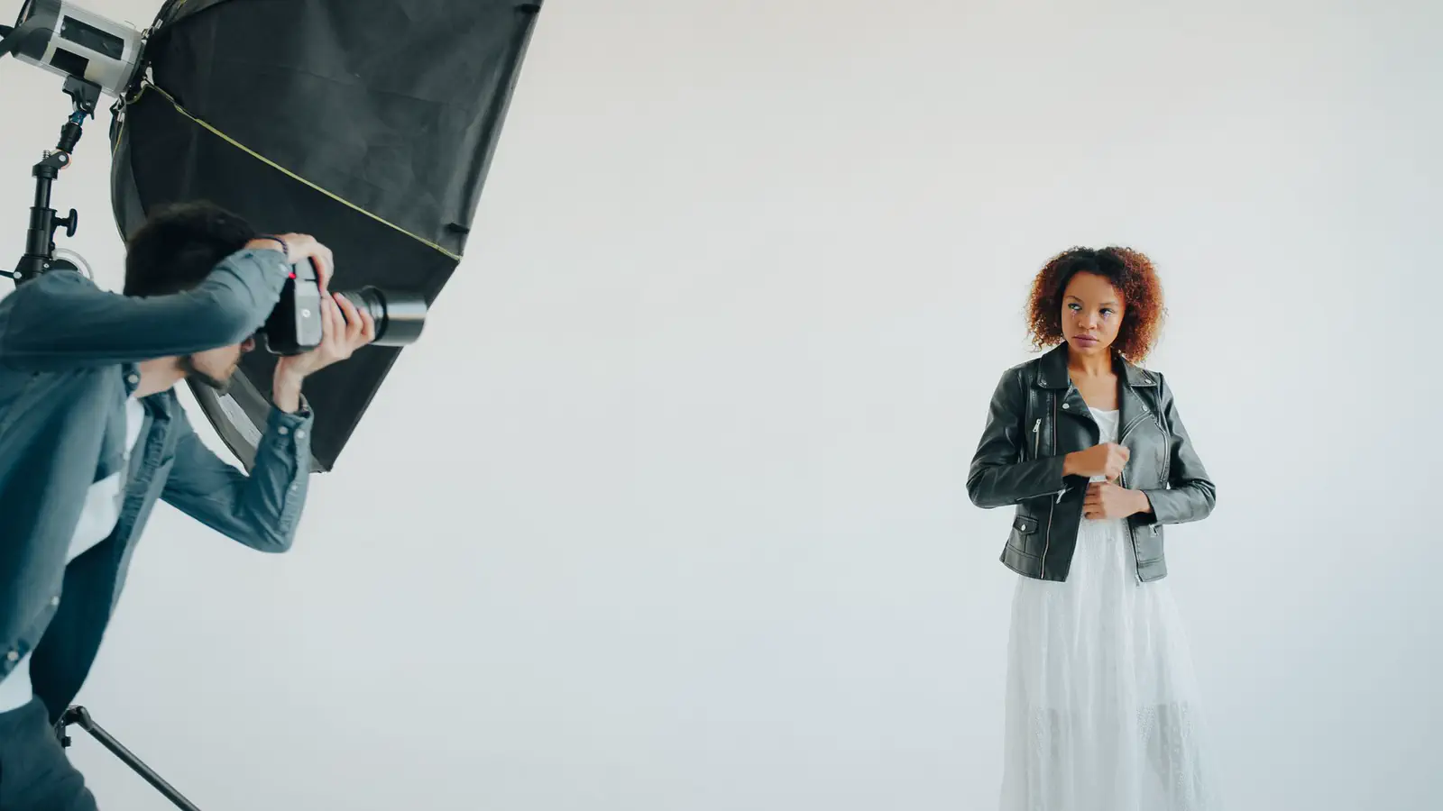 A photographer shooting a woman in a leather jacket and white skirt against a white wall.