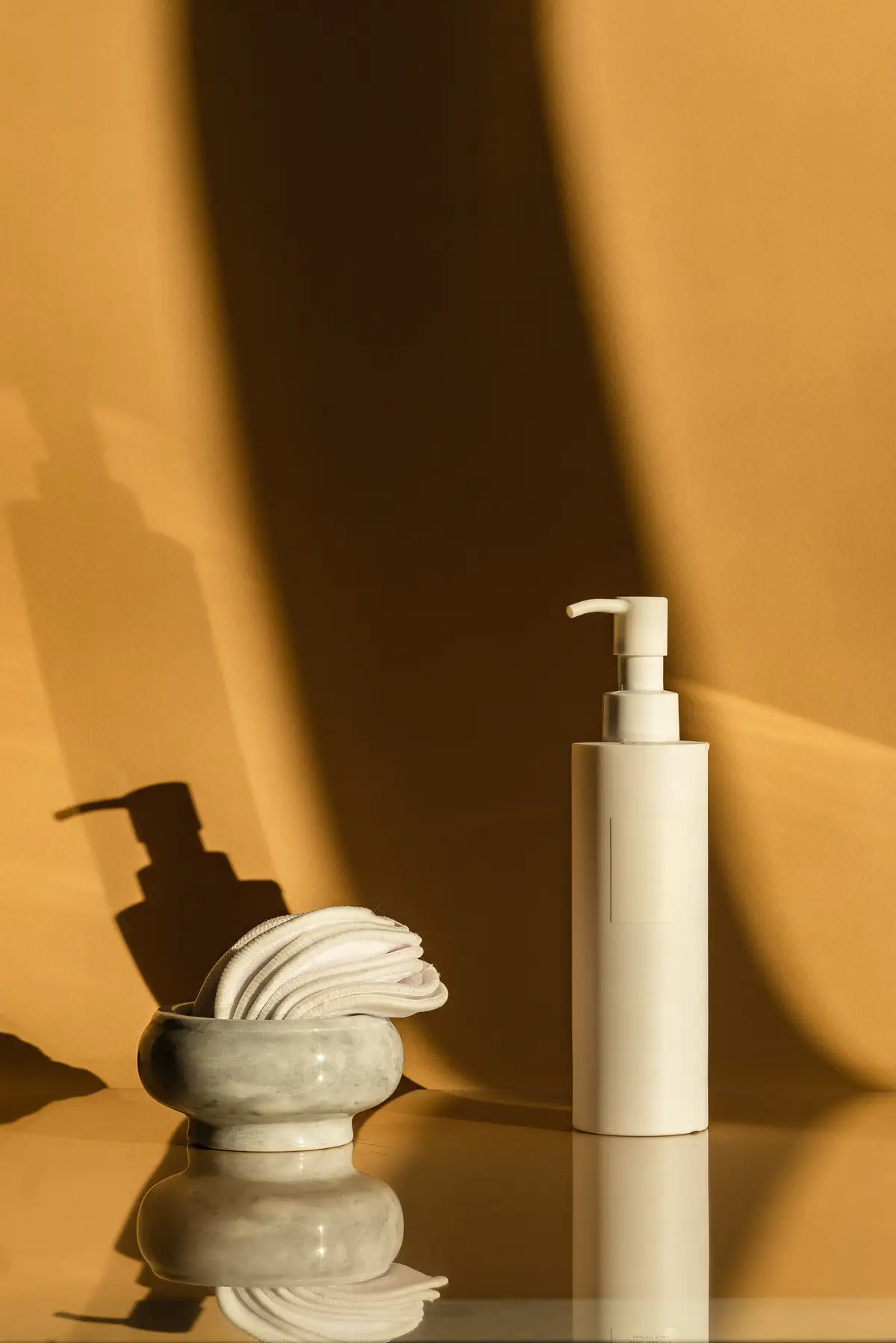The bottle beside a stone dish of folded cotton pads on amber cloth.