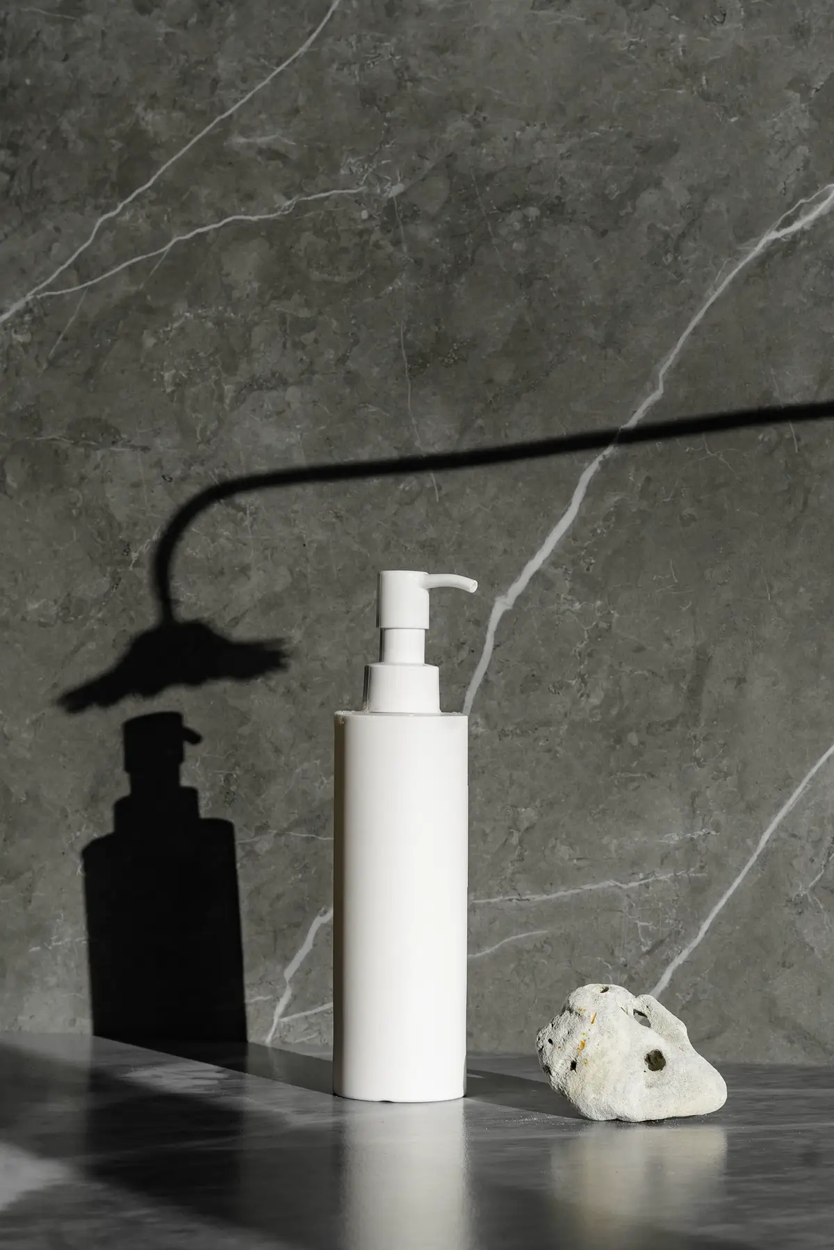 The bottle and a white pebble on grey marble under a flower-shaped shadow.