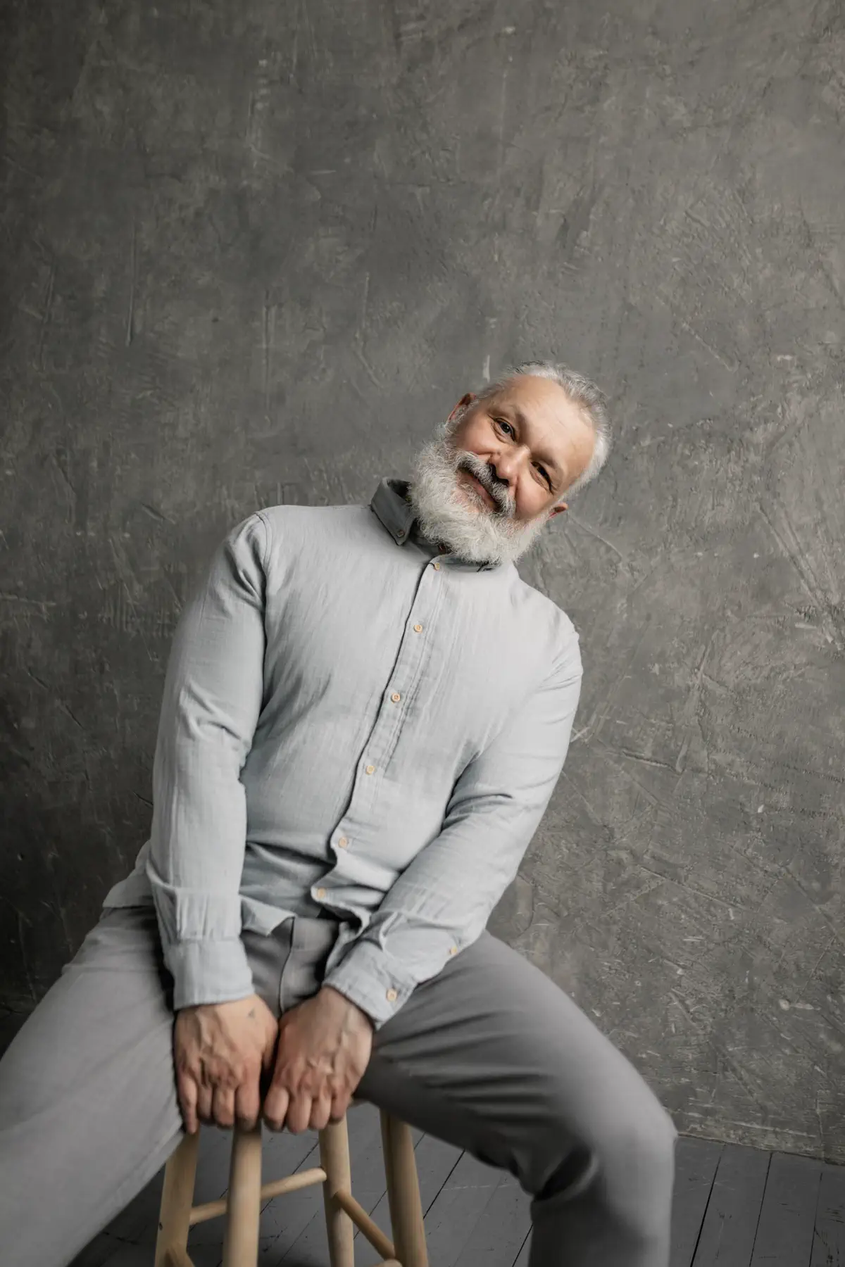 The bearded man on a wooden stool, tilting his head and laughing against a mottled grey backdrop.
