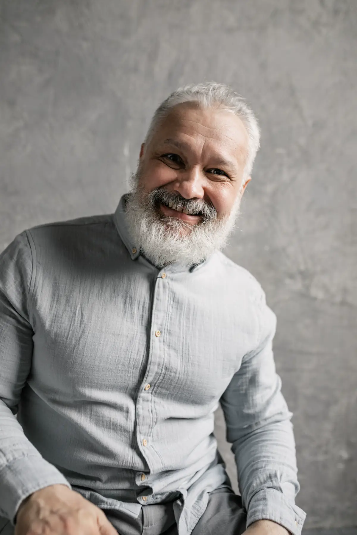 The bearded man in a grey shirt smiling warmly, hands resting on his knees.