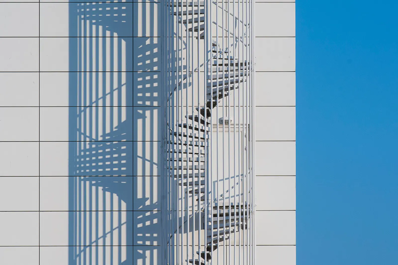 A spiral staircase casting a lattice shadow across a white tiled facade beside blue sky.