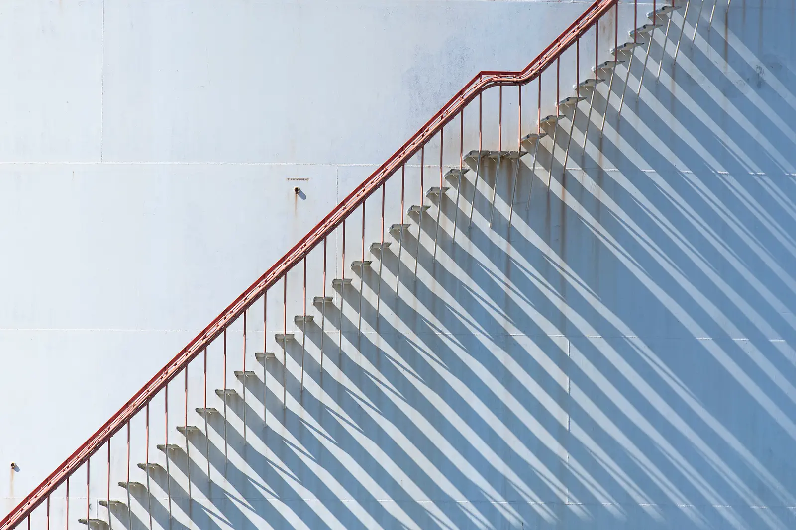 A red-railed stair climbing a white tank, its shadow repeating below.