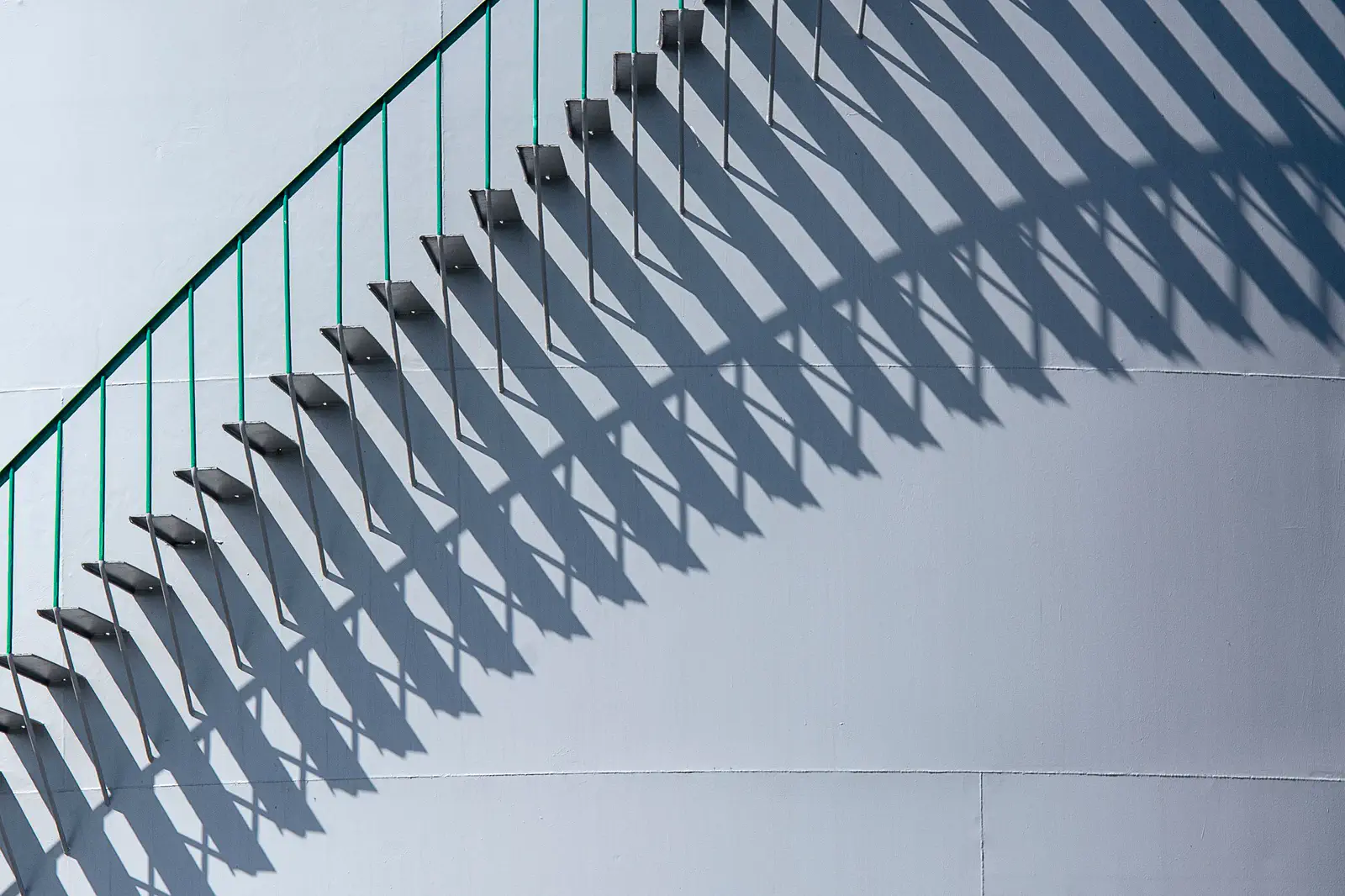A green-railed stair and its long shadow on a white curved wall.