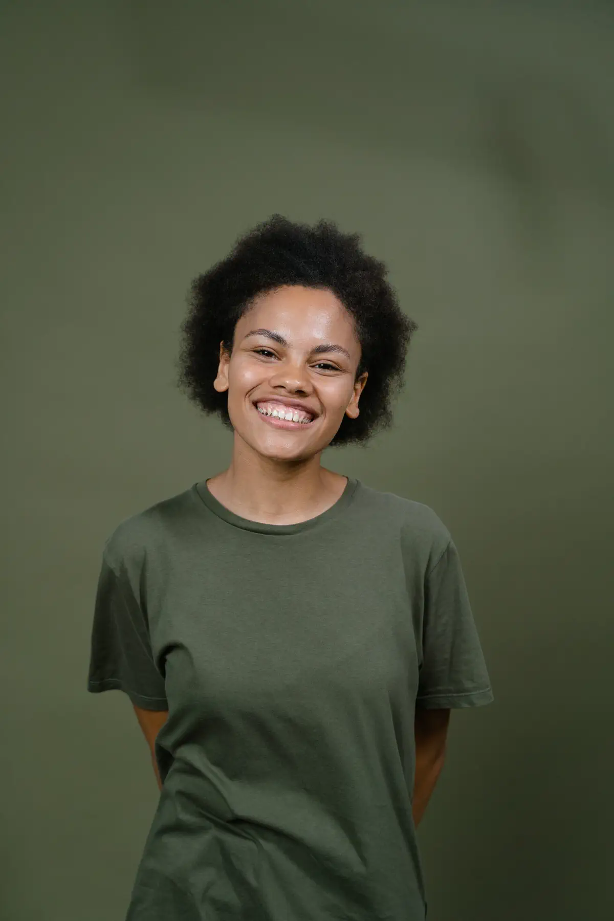 Studio portrait of a smiling young woman with natural curly hair in an olive T-shirt, hands behind her back, against an olive-green backdrop.