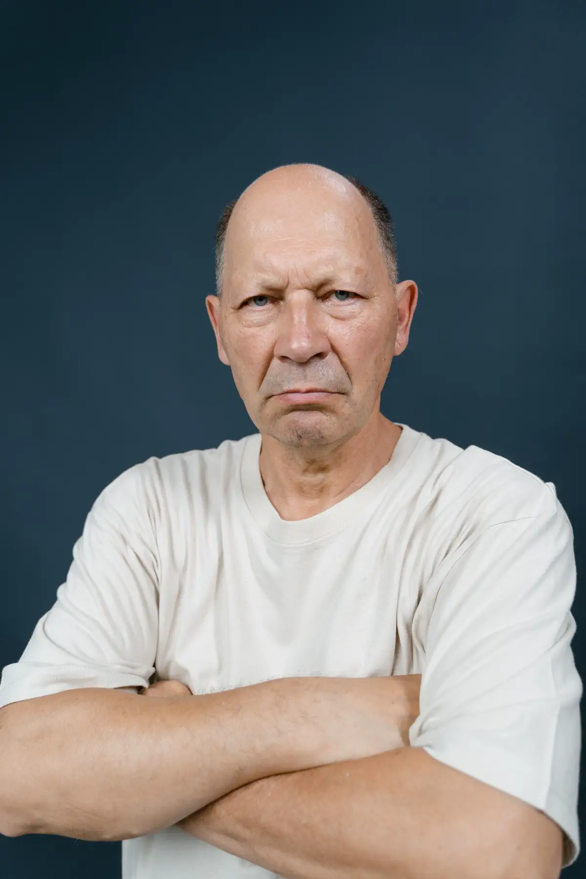 Bald older man in a white T-shirt with his arms crossed, frowning slightly, against a slate-blue backdrop.