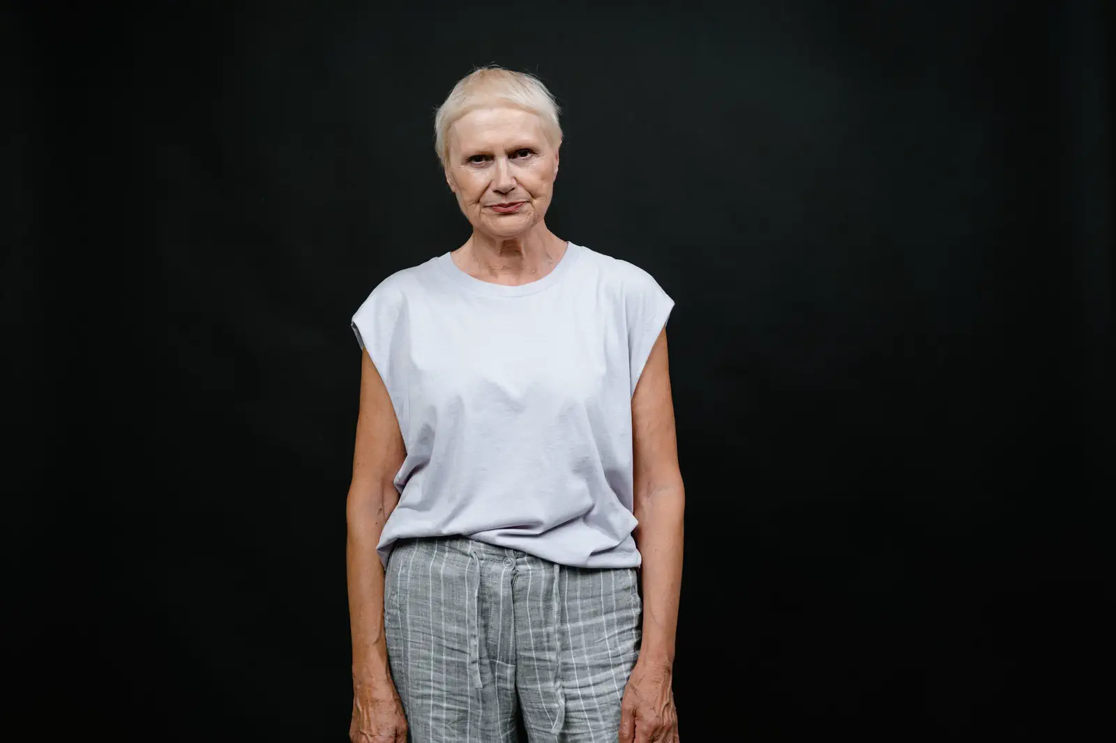 Full-length portrait of an older woman in a sleeveless top and checked trousers against a black backdrop.