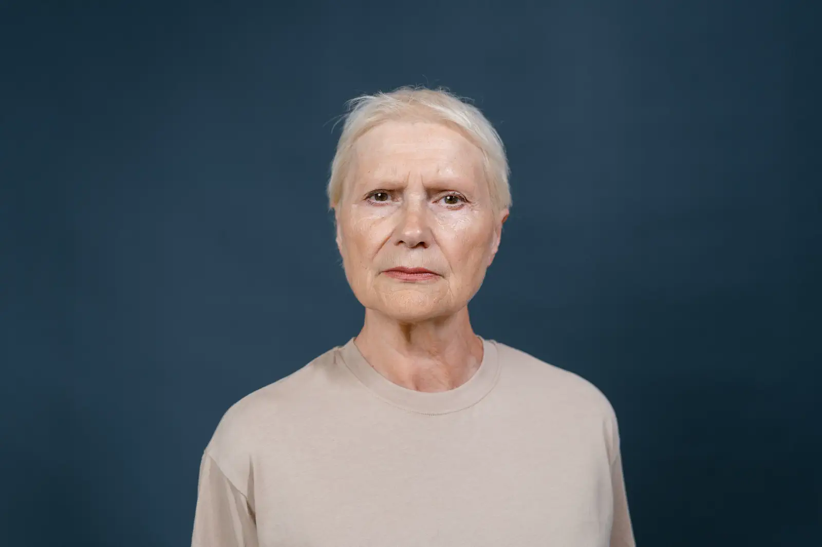 Older woman with short white hair in a beige T-shirt, chin raised, against a slate-blue backdrop.
