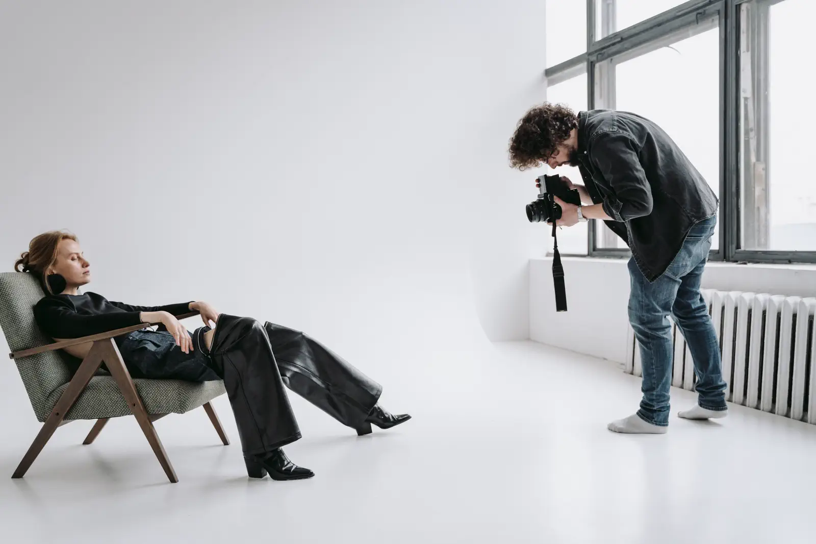 A sitter lounging in an armchair while the photographer bends to shoot beside a tall window and radiator.